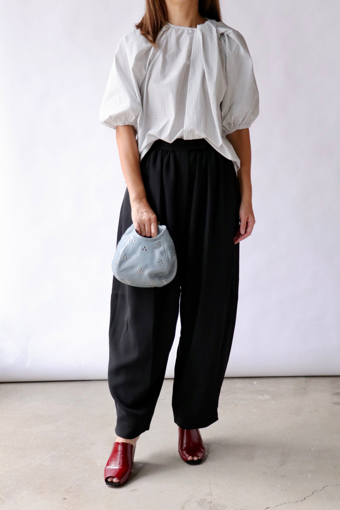 A model is shown from the shoulders down, wearing a white puff-sleeve blouse, loose black wide-leg trousers, and red shoes, while holding a small light blue pouch bag against a plain studio background.